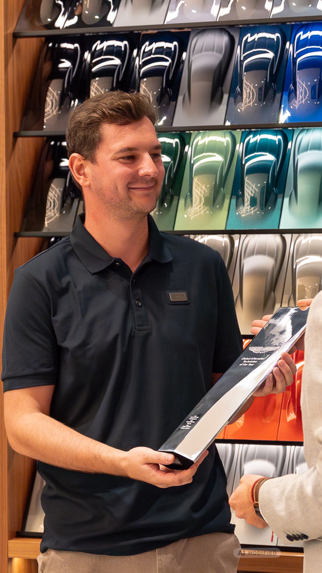 Felix Martini mit seiner Auszeichnung „Global Aftersales Technician of the Year“ vor einer Farbmusterwand im Aston Martin Showroom