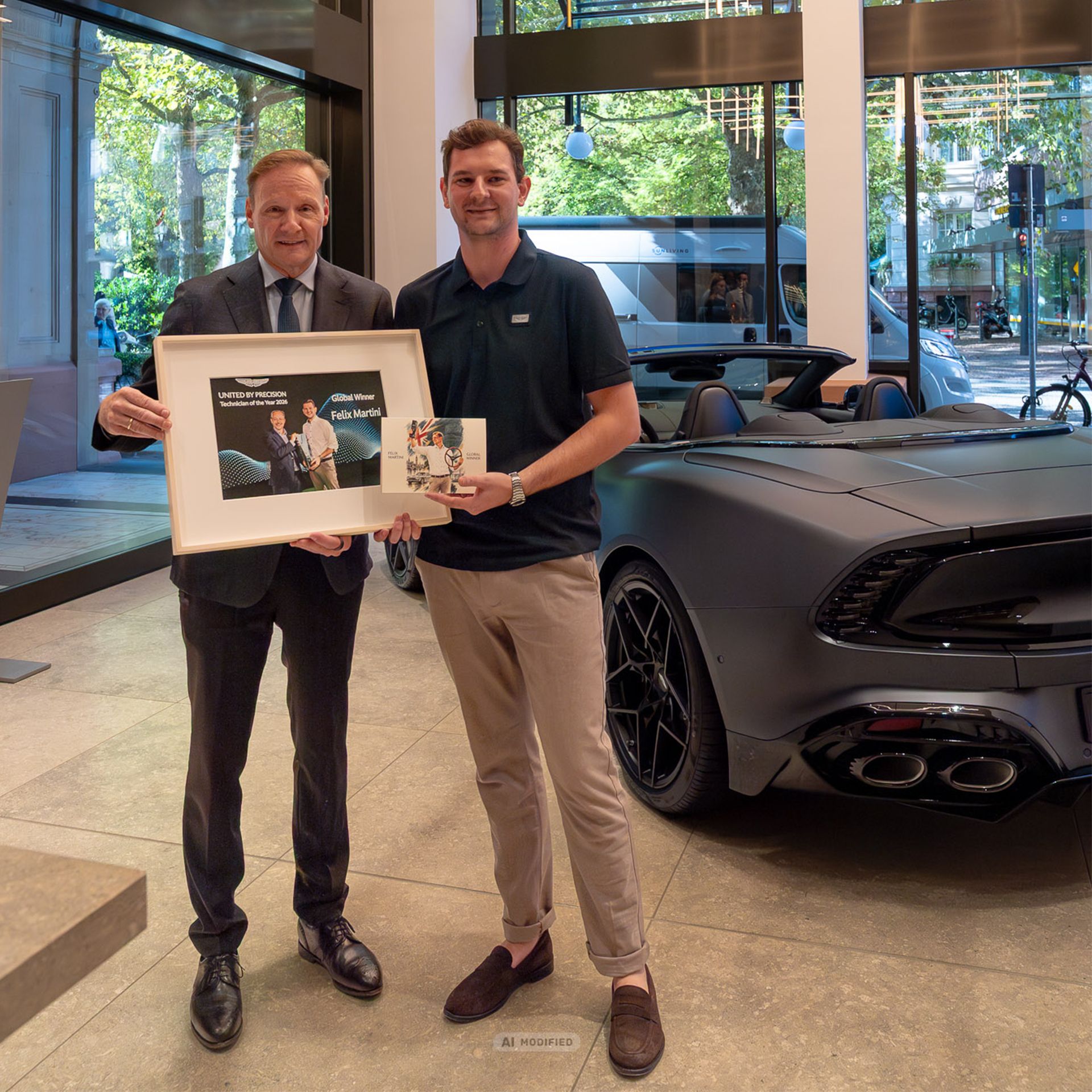 Felix Martini (rechts) mit gerahmter Urkunde „Technician of the Year 2026 – Global Winner“ im Aston Martin Showroom, daneben ein Aston Martin Cabrio