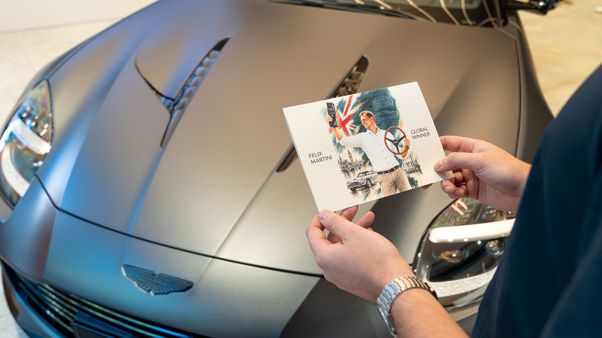 Illustrierte Karte „Felix Martini – Global Winner“ vor der Motorhaube eines Aston Martin